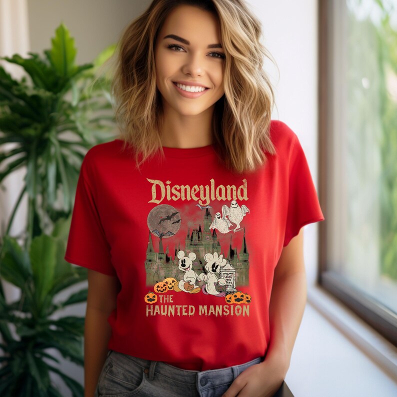 May include: Red t-shirt with a vintage-style graphic of Disneyland's Haunted Mansion. The graphic features Mickey Mouse and ghosts in front of the Haunted Mansion, with the text "Disneyland" and "The Haunted Mansion" above and below the image.