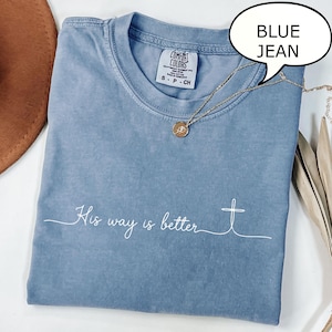 May include: A blue jean colored t-shirt with the words "His way is better" and a cross graphic in white. The shirt has a crew neck and a small tag at the neckline. A gold necklace with a pendant is draped on the shirt.