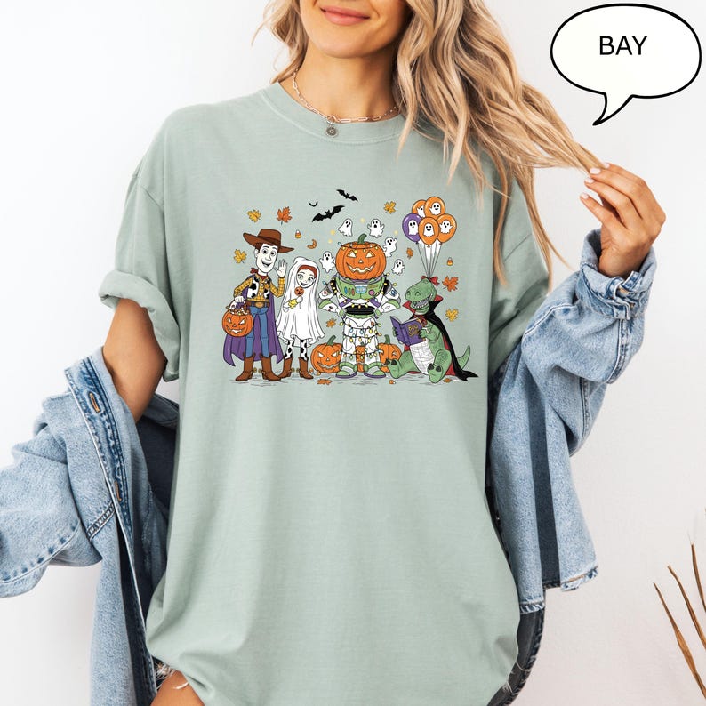 Puede incluir: Camiseta verde claro con un dise&ntilde;o de Halloween. El dise&ntilde;o muestra personajes de dibujos animados disfrazados: Woody, un fantasma, Buzz Lightyear y un dinosaurio. Las calabazas, los murci&eacute;lagos y las hojas rodean a los personajes.