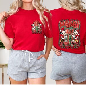 May include: Red t-shirt with a Christmas design featuring Mickey and Minnie Mouse. The front has a small pocket design, while the back displays "Merry Christmas" with the characters. The shirt is short-sleeved.