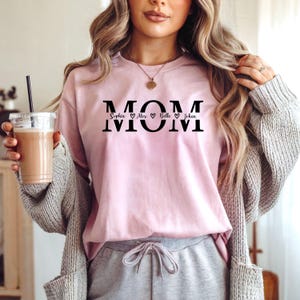 May include: A light pink sweatshirt with the word "MOM" in black block letters. Below the word "MOM" are the names Sophia, Alex, Belle, and Johan, each separated by a heart symbol. The sweatshirt is a casual style.