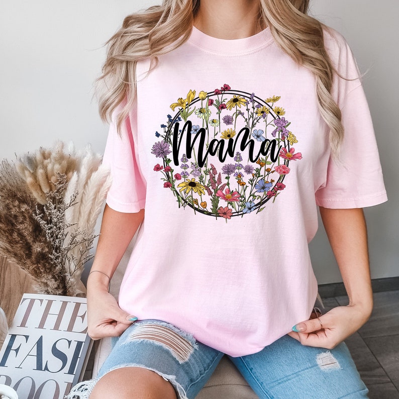 Peut inclure: T-shirt rose avec un motif de couronne de fleurs et le mot "Mama" en lettres noires.