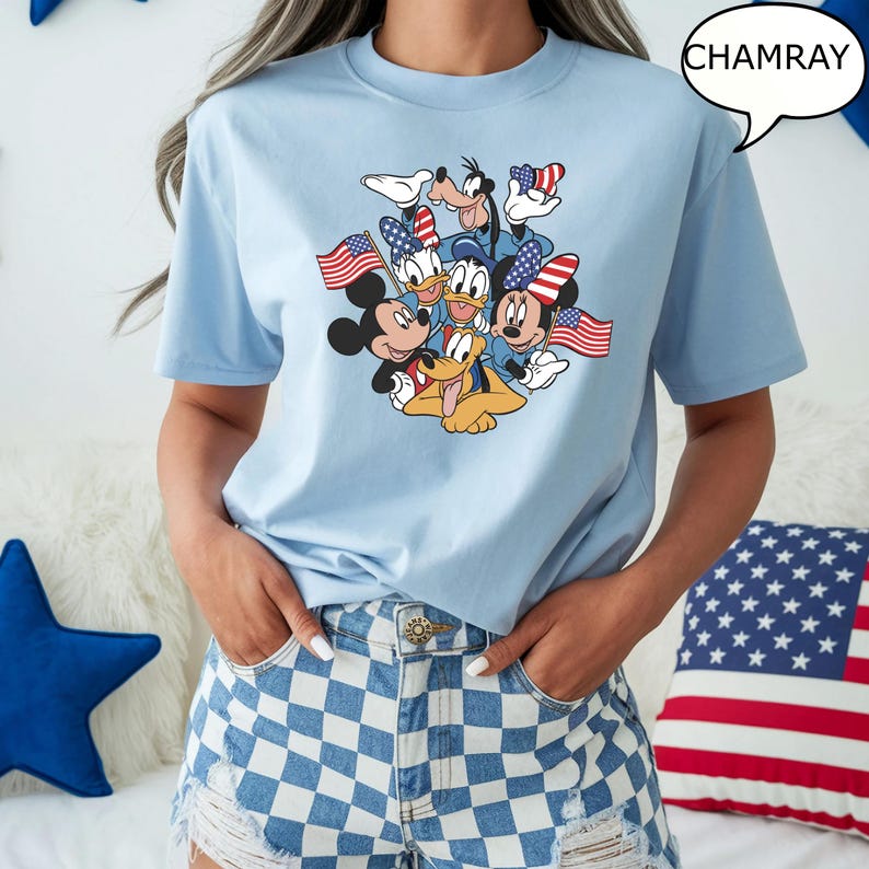 May include: Light blue t-shirt featuring a graphic of Mickey Mouse, Minnie Mouse, Donald Duck, Daisy Duck, Goofy, and Pluto holding American flags. The shirt has a crew neck and short sleeves. The word "CHAMRAY" is in a speech bubble.