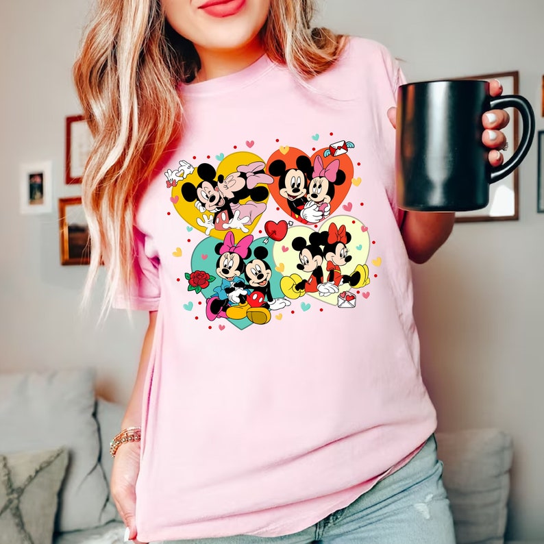Puede incluir: Camiseta rosa con un gr&aacute;fico de Mickey Mouse y Minnie Mouse en cuatro poses diferentes, cada una dentro de una forma de coraz&oacute;n. Los corazones est&aacute;n rodeados de corazones peque&ntilde;os y otros elementos decorativos.