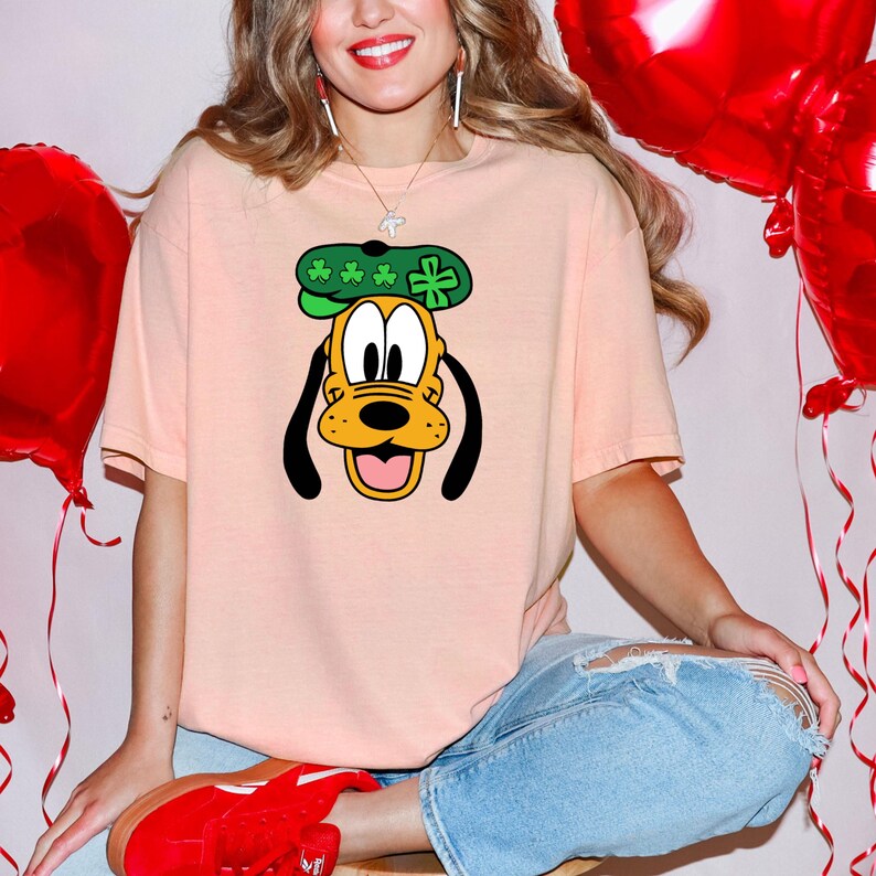 Puede incluir: Camiseta de color melocot&oacute;n claro con una ilustraci&oacute;n de dibujos animados de Pluto con un sombrero verde con tr&eacute;boles. La cara de Pluto es amarilla con orejas negras y una lengua rosa. La camiseta se combina con vaqueros azules y zapatos rojos.