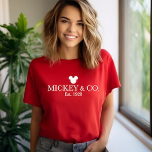 Puede incluir: Camiseta roja con una cabeza de Mickey Mouse blanca y el texto "MICKEY & CO. Est. 1928"