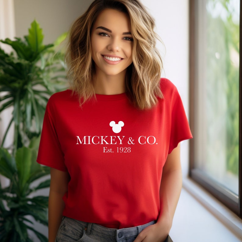 Puede incluir: Camiseta roja con una cabeza de Mickey Mouse blanca y el texto "MICKEY & CO. Est. 1928"