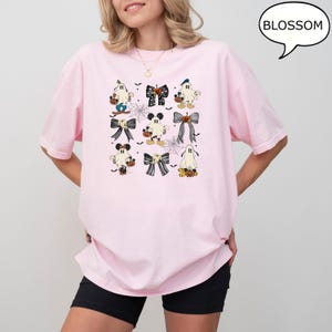 Puede incluir: Una camiseta rosa claro con un dise&ntilde;o de Halloween. El dise&ntilde;o incluye fantasmas de dibujos animados, lazos y personajes inspirados en Mickey Mouse, todos con un dise&ntilde;o espeluznante. La palabra "BLOSSOM" est&aacute; en un bocadillo.