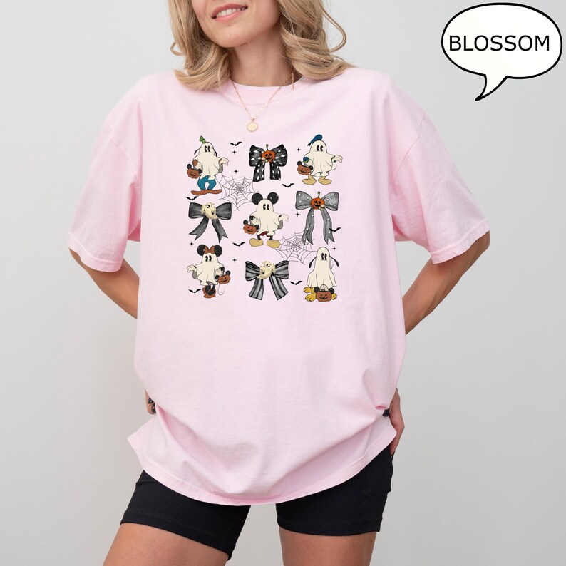 Puede incluir: Una camiseta rosa claro con un dise&ntilde;o de Halloween. El dise&ntilde;o incluye fantasmas de dibujos animados, lazos y personajes inspirados en Mickey Mouse, todos con un dise&ntilde;o espeluznante. La palabra "BLOSSOM" est&aacute; en un bocadillo.