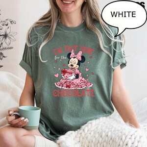Puede incluir: Camiseta verde con un gr&aacute;fico de Minnie Mouse. Minnie lleva un vestido rosa y sostiene una caja en forma de coraz&oacute;n. El texto "I'M JUST HERE for the CHOCOLATE" est&aacute; impreso en rojo.