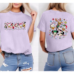 May include: A light purple t-shirt featuring a festive design with Disney characters, including Mickey Mouse, Donald Duck, and Goofy, wearing Christmas hats and surrounded by string lights. The back of the shirt also has a similar design.