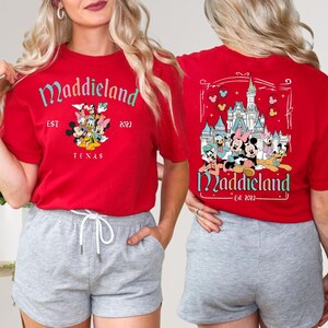 K&ouml;nnte beinhalten: Rotes T-Shirt mit einem Motiv von Mickey Mouse und seinen Freunden vor dem Schloss von Aschenputtel. Der Text "Maddieland Est 2023" ist auf dem T-Shirt aufgedruckt.