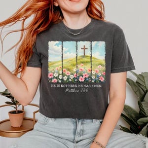 May include: A black t-shirt featuring a graphic of a cross on a hill with flowers. The text "He is not here. He has risen. Matthew 28:6" is printed below the graphic.
