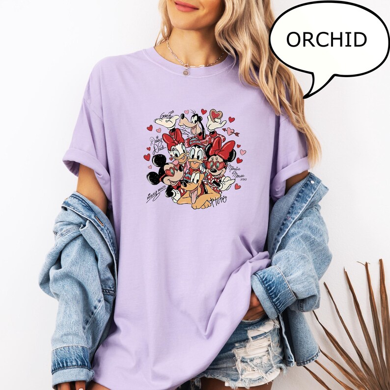 May include: Orchid-coloured t-shirt featuring a graphic of Disney characters, including Mickey and Minnie Mouse, Donald Duck, and Pluto, surrounded by hearts. The word "ORCHID" is in a speech bubble. The shirt has short sleeves.