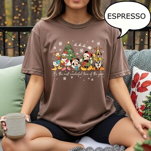 May include: Espresso-colored t-shirt featuring a festive Disney character design. The graphic includes Mickey Mouse, Donald Duck, and other characters, along with a Christmas tree and snowflakes. The text reads, "It's the most wonderful time of the year."