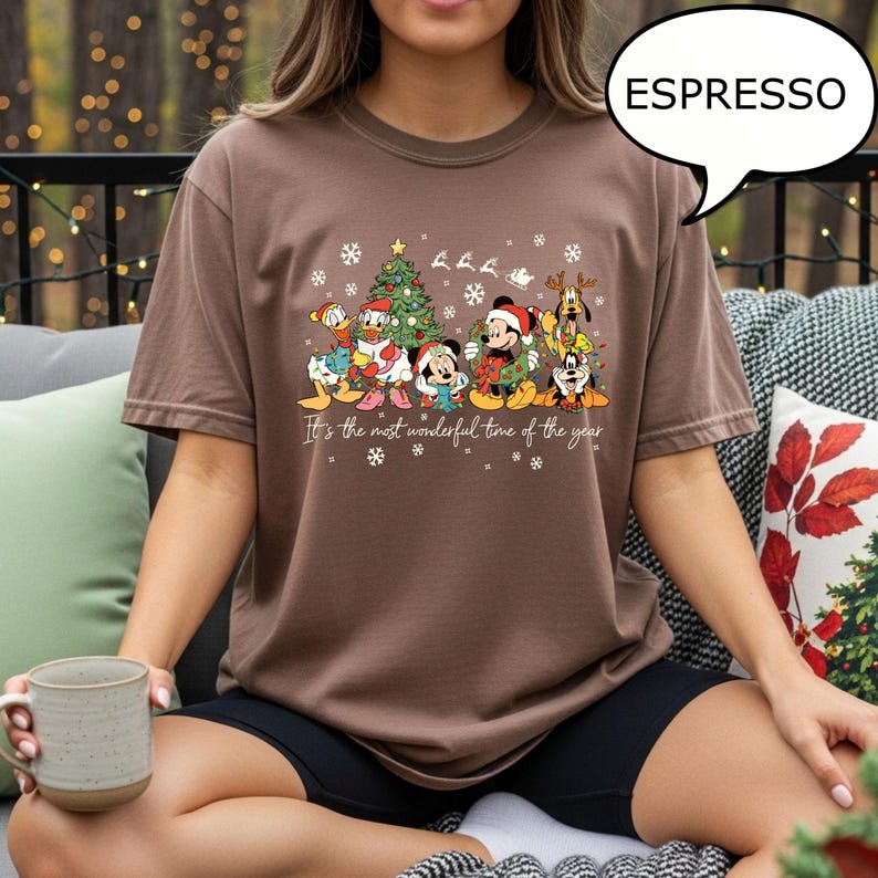 May include: Espresso-colored t-shirt featuring a festive Disney character design. The graphic includes Mickey Mouse, Donald Duck, and other characters, along with a Christmas tree and snowflakes. The text reads, "It's the most wonderful time of the year."