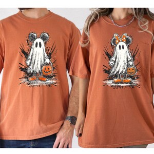 May include: Two orange t-shirts featuring a cartoon ghost with Mickey Mouse ears and a pumpkin bucket. The ghost is wearing orange shoes and is set against a black splatter design. One shirt has a bow on the ears.