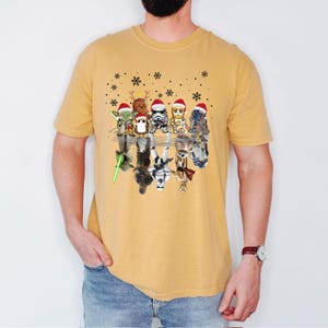 Puede incluir: Una camiseta amarilla con un gr&aacute;fico de Star Wars con tem&aacute;tica navide&ntilde;a. El gr&aacute;fico presenta un grupo de personajes de Star Wars con gorros de Pap&aacute; Noel, reflejados en un charco de agua. Los personajes incluyen a Yoda, Chewbacca, Darth Vader, R2-D2 y C-3PO.