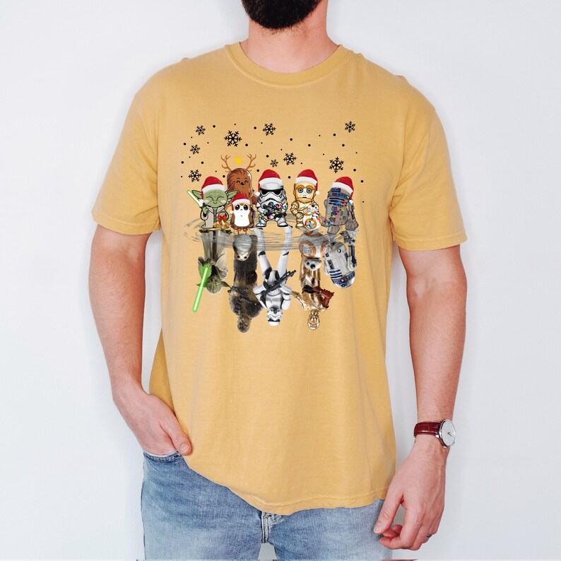 Puede incluir: Una camiseta amarilla con un gr&aacute;fico de Star Wars con tem&aacute;tica navide&ntilde;a. El gr&aacute;fico presenta un grupo de personajes de Star Wars con gorros de Pap&aacute; Noel, reflejados en un charco de agua. Los personajes incluyen a Yoda, Chewbacca, Darth Vader, R2-D2 y C-3PO.