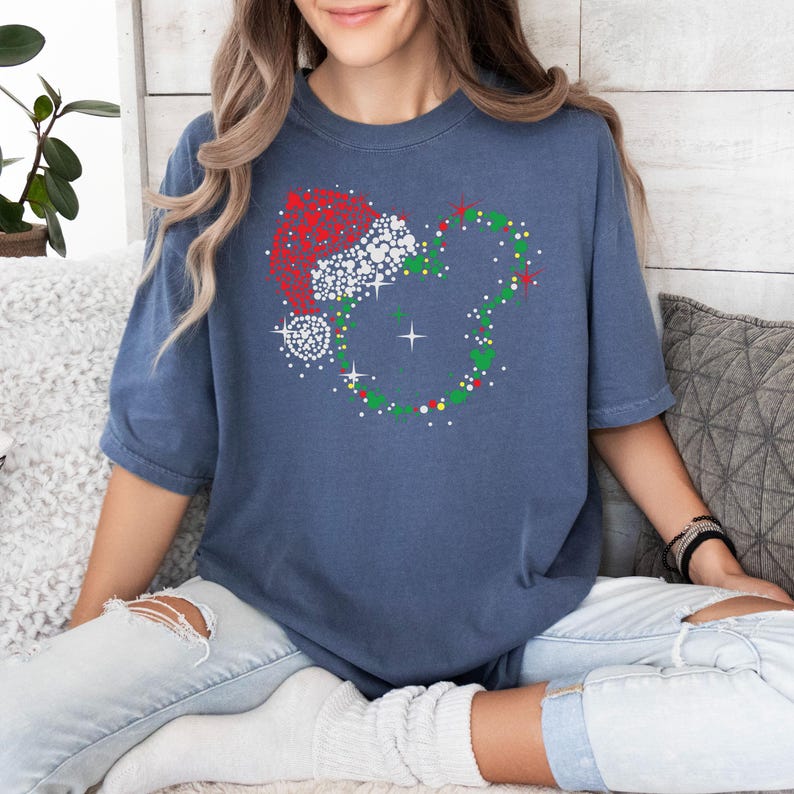 Puede incluir: Una camiseta azul con un dise&ntilde;o navide&ntilde;o. El gr&aacute;fico combina un gorro de Pap&aacute; Noel con una forma de cabeza de Mickey Mouse, adornada con detalles rojos, blancos, verdes y amarillos. La camiseta es de estilo casual.