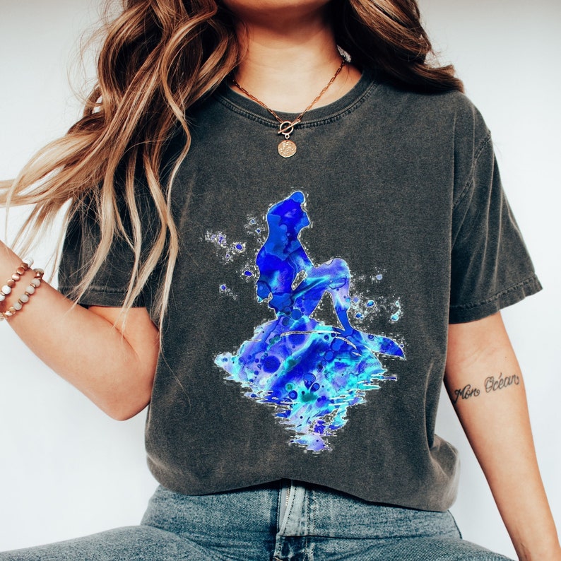 Puede incluir: Una camiseta gris oscuro con una ilustraci&oacute;n de acuarela azul de una sirena sentada en una roca. La sirena est&aacute; delineada en negro y tiene una larga cola fluida. El fondo es una lavada de acuarela azul claro.