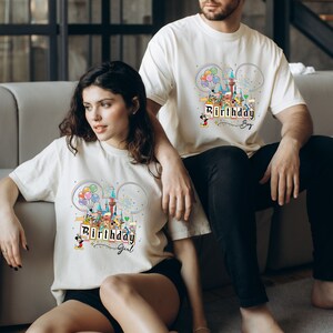 Puede incluir: Dos camisetas blancas con un gr&aacute;fico colorido de un castillo, globos y fuegos artificiales. El texto "Birthday Boy" y "Birthday Girl" est&aacute; en las camisetas.