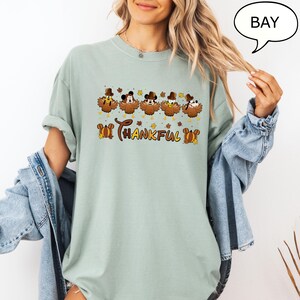 May include: A light green t-shirt featuring a Thanksgiving-themed graphic. The design includes cartoon turkey characters with Mickey Mouse ears and the word "Thankful" in a decorative font. The shirt has short sleeves and a relaxed fit.