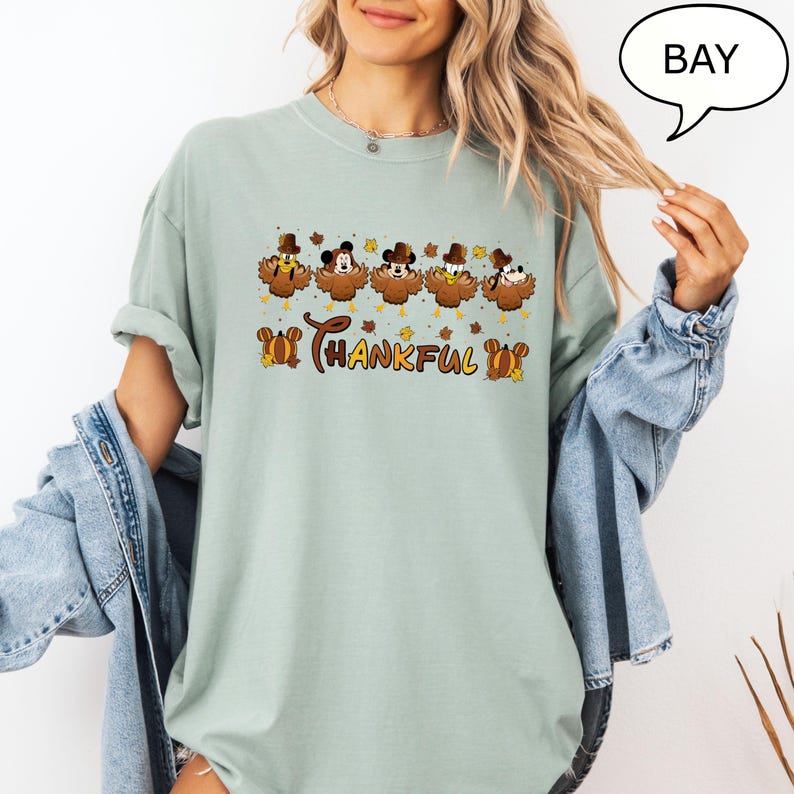 May include: A light green t-shirt featuring a Thanksgiving-themed graphic. The design includes cartoon turkey characters with Mickey Mouse ears and the word "Thankful" in a decorative font. The shirt has short sleeves and a relaxed fit.