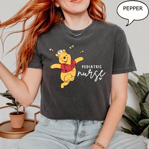May include: Dark gray t-shirt featuring a cartoon Winnie the Pooh character wearing a nurse's hat and red vest, with the words "Pediatric Nurse" in white script. The design includes bees. The shirt is a comfortable fit.