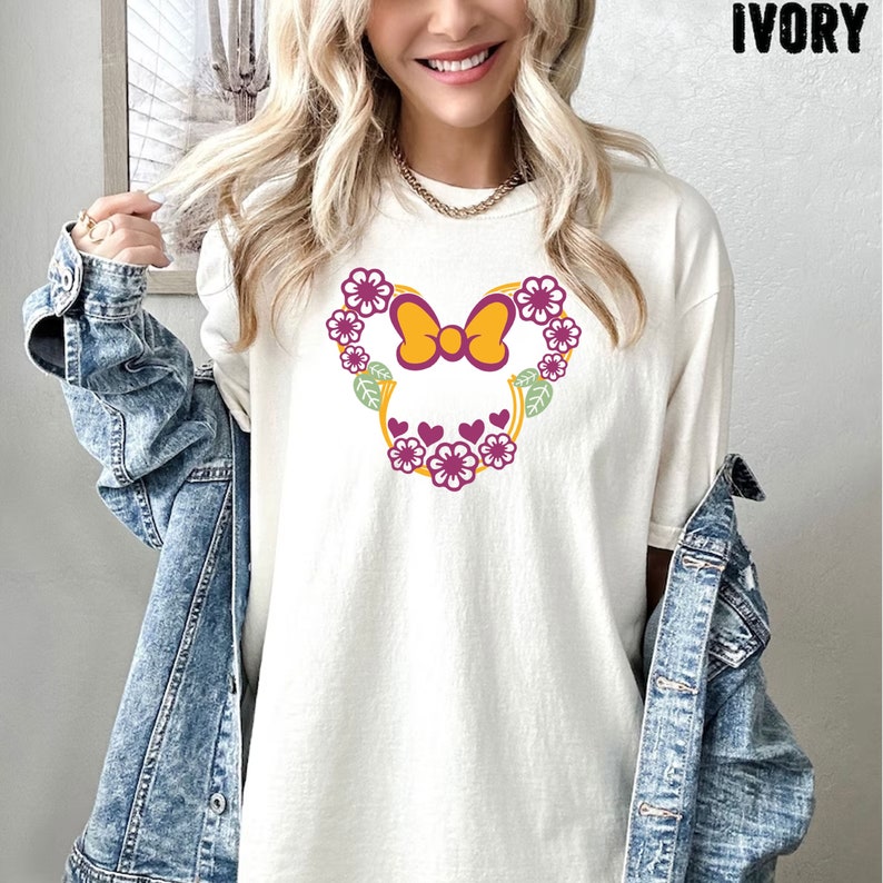 May include: White t-shirt with a floral design in the shape of a mouse head. The flowers are purple and yellow with green leaves. The bow is yellow with a purple center.