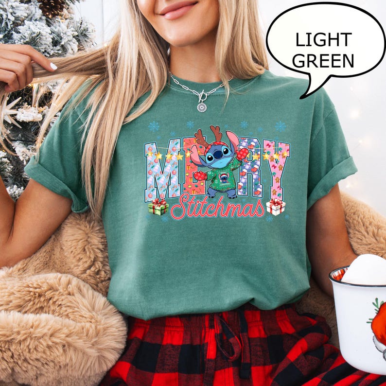 Op de afbeelding: Lichtgroen T-shirt met de tekst "MERRY Stitchmas" en een stripfiguur met een rendiergeweige haarband en een groene trui. Het shirt heeft een ronde hals en korte mouwen. De tekst is in een kleurrijk, feestelijk lettertype.