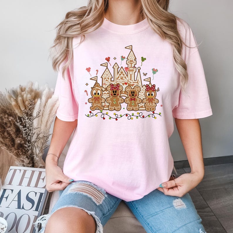 May include: A light pink t-shirt featuring a gingerbread castle and gingerbread characters. The gingerbread castle has flags and is decorated with candy canes and hearts. The gingerbread characters are wearing bows and bow ties. A string of colorful lights is at the bottom.