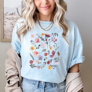 May include: A light blue t-shirt with a floral print featuring a variety of flowers in shades of pink, red, yellow, and purple.