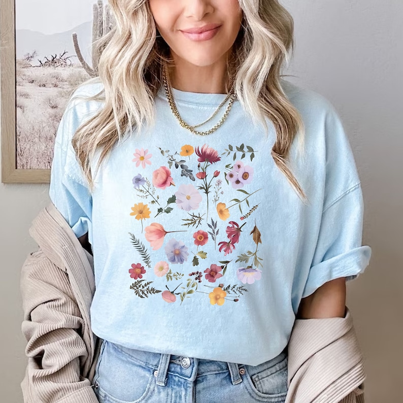 May include: A light blue t-shirt with a floral print featuring a variety of flowers in shades of pink, red, yellow, and purple.