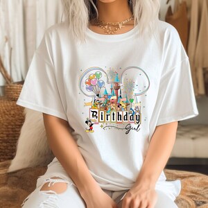 Puede incluir: Camiseta blanca con un gr&aacute;fico colorido de Mickey Mouse y sus amigos en un parque tem&aacute;tico de Disney. El gr&aacute;fico incluye un castillo, globos, fuegos artificiales y el texto "Birthday Girl".