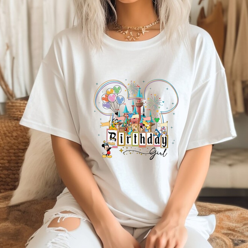 Puede incluir: Camiseta blanca con un gr&aacute;fico colorido de Mickey Mouse y sus amigos en un parque tem&aacute;tico de Disney. El gr&aacute;fico incluye un castillo, globos, fuegos artificiales y el texto "Birthday Girl".