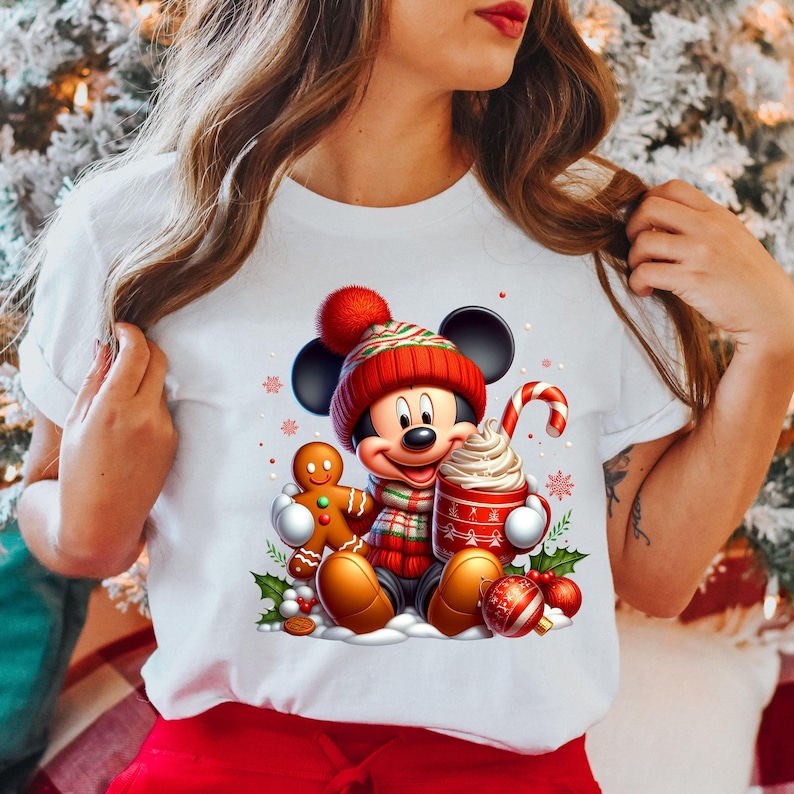Puede incluir: Camiseta blanca con un gr&aacute;fico navide&ntilde;o que presenta a Mickey Mouse con un gorro de Pap&aacute; Noel rojo y blanco, sosteniendo una taza de chocolate caliente con crema batida y un bast&oacute;n de caramelo, y un hombre de jengibre.