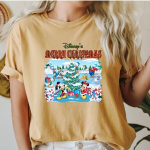 May include: A yellow t-shirt with a Christmas scene featuring Mickey Mouse and other Disney characters. The text "Disney's Merry Christmas" is printed on the shirt.