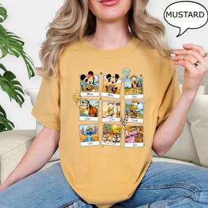 Puede incluir: Una camiseta amarilla mostaza con un estampado gr&aacute;fico de personajes de Disney con atuendos de safari. Los personajes incluyen a Mickey Mouse, Minnie Mouse, Pluto, Goofy, Donald Duck, Daisy Duck, Stitch, Chip y Dale. El texto "MUSTARD" est&aacute; en un bocadillo de di&aacute;logo en la esquina superior derecha de la imagen.