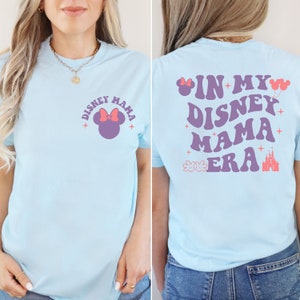May include: Pale blue t-shirt featuring the text "DISNEY MAMA" and a Minnie Mouse design on the front. The back of the shirt displays "IN MY DISNEY MAMA ERA" with Disney-themed graphics.