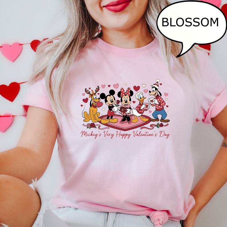 Puede incluir: Camiseta rosa claro con un dise&ntilde;o de San Valent&iacute;n que presenta a los personajes de Disney Mickey, Minnie, Donald, Goofy y Pluto. La frase "Mickey's Very Happy Valentine's Day" est&aacute; impresa debajo de los personajes. Tambi&eacute;n es visible un bocadillo con la palabra "BLOSSOM".