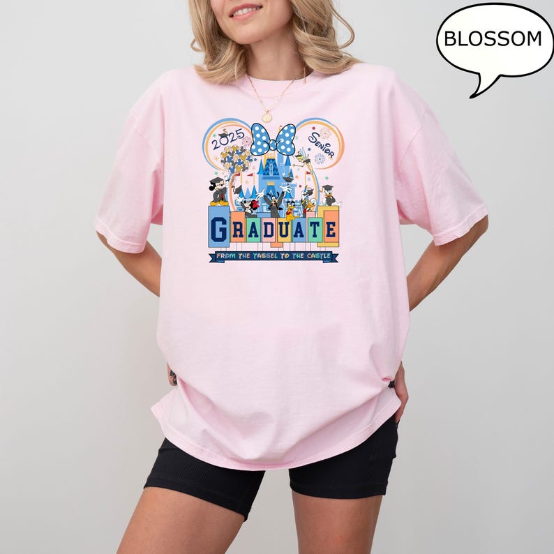 以下が含まれることがあります： シンデレラ城の前でミッキーマウスとその仲間たちのグラフィックが描かれたピンクのTシャツ。Tシャツには「2025 Senior Graduate From the Tagel to the Castle」の文字がプリントされています。