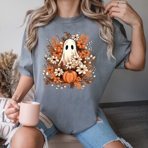 Op de afbeelding: Een grijs T-shirt met een witte geest die een oranje pompoen vasthoudt, omringd door bruine en witte bloemen. De geest heeft twee zwarte ogen.