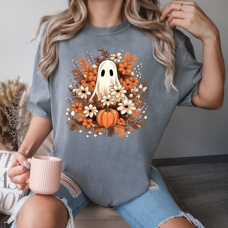 Op de afbeelding: Een grijs T-shirt met een witte geest die een oranje pompoen vasthoudt, omringd door bruine en witte bloemen. De geest heeft twee zwarte ogen.