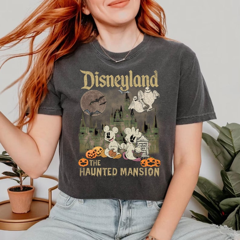 May include: A black t-shirt with a vintage-style graphic of Disneyland's Haunted Mansion. The graphic features Mickey Mouse and a ghost holding jack-o'-lanterns in front of the Haunted Mansion.