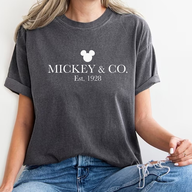 Puede incluir: Una camiseta gris oscuro con una cabeza de Mickey Mouse blanca y el texto "MICKEY & CO. Est. 1928"