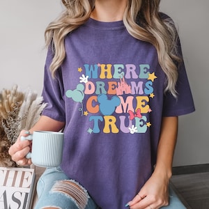 May include: Purple t-shirt with the text "Where Dreams Come True" in colorful letters. The design includes a castle, Mickey Mouse ears, stars, and a bow.