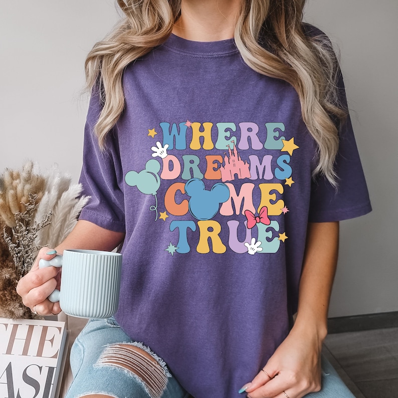 May include: Purple t-shirt with the text "Where Dreams Come True" in colorful letters. The design includes a castle, Mickey Mouse ears, stars, and a bow.
