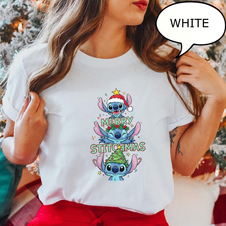 May include: White t-shirt featuring a stacked design of three blue cartoon characters, each adorned with Christmas-themed elements. The text "MERRY STITCHMAS" is displayed in green and red. A speech bubble above the shirt says "WHITE".