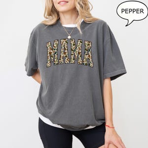 May include: A gray oversized t-shirt with a camo print "MAMA" graphic. The model is wearing black shorts and a silver necklace.